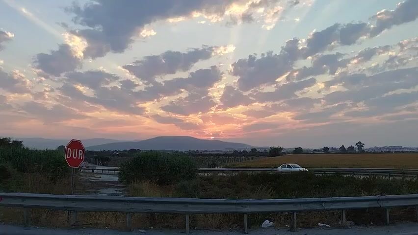 A stop sign reading "DUR" is visible on the left side of the road. A white car drives past on the right, with a sunset in the background. The sun is partially obscured by clouds, casting rays across the sky. Hills are visible in the distance.