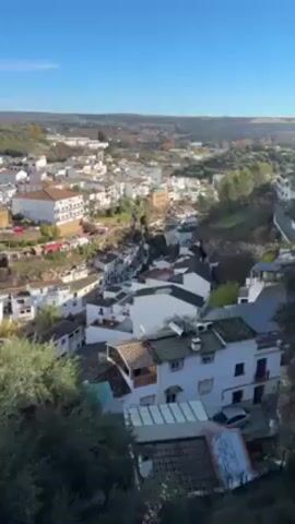 The video shows a high-angle panoramic view of a town nestled in a hilly landscape, likely somewhere in southern Europe. The town is predominantly white, with buildings clustered closely together, and some structures that appear to be built into or onto the natural rock formations. Some buildings have red tiled roofs. Olive groves cover the hills, extending into the distance. The sky is a clear, bright blue with a few wispy clouds. The camera pans slightly to the right, providing a wider view of the town and the surrounding landscape.