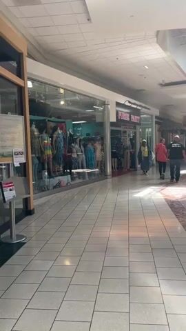 The video shows a view inside a shopping mall hallway. On the left, there is a store window display featuring mannequins dressed in colorful clothing, including dresses and what appears to be resort wear. The store name "PURSE PARTY" is visible above the store next to it. A woman wearing a blue outfit and a yellow scarf is walking towards the camera from the direction of the "PURSE PARTY" store. Behind her, a woman in a pink shirt and a child are walking in the same direction. Further down the hallway, a man in a black shirt is walking away from the camera. The hallway has tiled floors and fluorescent lighting. Sunlight is streaming in from the right side of the hallway, casting shadows on the floor.