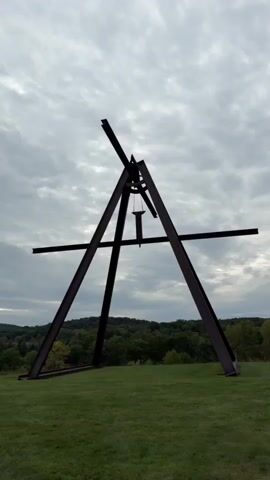 The video shows a large, dark metal sculpture in the shape of an A-frame with a horizontal beam crossing the top. A smaller metal object hangs from a chain in the center of the A-frame. The sculpture is situated on a grassy hill with trees and rolling hills in the background. The sky is overcast with clouds. The camera is positioned low, looking up at the sculpture. There is no visible activity or interaction.