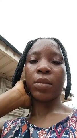 A young woman with braided hair is seen in a video, with her face close to the camera. She moves her hands to her neck and face as she looks around. The background appears to be outdoors, with a building and some vegetation visible. The woman is wearing a patterned top.