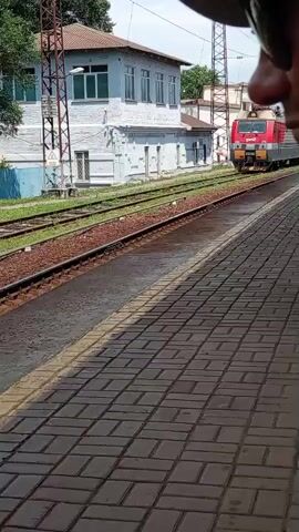 A train approaches a station platform. The train is red and silver. There is a building behind the tracks. The platform is made of brick. The person filming is at the station.