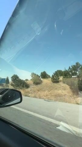 A passenger's bare foot is visible resting on the dashboard of a car. The car is in motion, evidenced by the blurred scenery outside the window. A road sign is visible through the side window, listing distances to Afyonkarahisar, Uşak, and Antalya. The sky is clear and blue, with some sparse vegetation and trees lining the roadside. The interior of the car is partially visible, including part of the dashboard and the car's interior pillar.