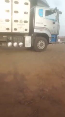 The video depicts a truck with the name "Dangote" on the side in an open lot. It is a gray truck with blue accents. The truck has a SINOTRUK logo, and other writing includes "DSR-CNG-1F-030" and "KZR-906XC." The truck appears to be stationary in the opening shot, but then it moves forward. The camera pans to show the area around the truck, which includes other trucks and some buildings in the background. The ground appears to be red dirt. A person on a motorcycle briefly passes in front of the camera.