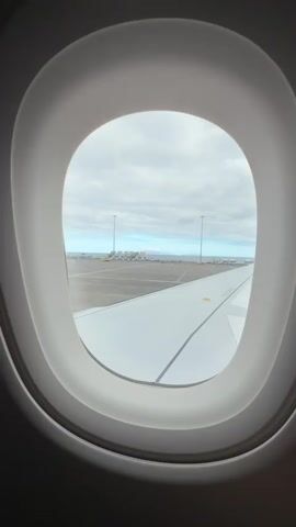 Here's a description of the video:
The video shows a view out of an airplane window. The plane is on the ground at an airport. The wing of the plane is visible below the window. Beyond the wing, the airport tarmac can be seen with some airport vehicles and equipment parked. In the distance, the ocean and an island are visible under a cloudy sky.