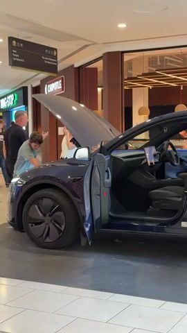 A dark blue Tesla Model Y is parked indoors, possibly in a mall, with its hood and driver's side door open. The interior of the car, including the seats and dashboard, is visible. A young man in a black hoodie is seated in the driver's seat, appearing to interact with something on the seat. Another person, an adult male, is standing near the open hood, looking into the engine compartment. In the background, a Chipotle restaurant is visible, and other people are walking around. The lighting suggests an indoor environment, likely during the day.