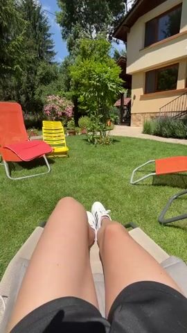 A person's legs and feet, wearing white sneakers and black shorts, are visible in the foreground, lying on a reclining chair. The chair is positioned on a lush green lawn in a backyard. Behind the person, in the middle ground, are three lounge chairs: one orange with a red cushion, one yellow, and another orange chair. A small tree and various potted plants are scattered across the lawn. In the background, a two-story house with a light-colored exterior and dark windows stands prominently. The house has a pitched roof and a balcony or porch area. Sunlight is casting shadows on the lawn, indicating a bright, sunny day. The sky above is blue with scattered white clouds. The overall scene depicts a relaxed outdoor setting, likely a backyard during the daytime.
