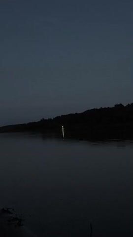 The video shows a dark, calm body of water with a wooded hillside in the background. The sky is a deep, dark blue, suggesting it is either dusk or dawn. In the center of the water, a single white object, possibly a buoy or a small boat, is visible. The object is illuminated by a light source, casting a reflection on the water's surface. The overall mood of the video is peaceful and serene, with a touch of mystery due to the darkness and the solitary illuminated object.