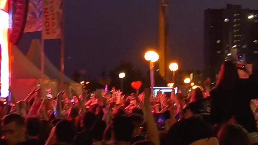 This video shows a crowded outdoor event at dusk. A large group of people are visible, many with their hands raised in the air. Some are holding up phones, likely to record the event or take pictures. In the background, there are tall buildings with lights on in the windows. There are also lights from the event itself, including streetlights and what appear to be stage lights. A Russian flag is visible on a flagpole. There are also tents and banners, suggesting a festival or concert. One person in the foreground is being carried on someone's shoulders, and they are holding up a phone. Another person is holding up a red heart-shaped balloon. The overall atmosphere appears to be festive and energetic.