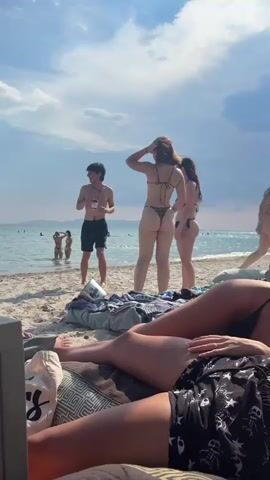 This is a short video clip taken on a sunny day at Fanari, Greece. The scene is a sandy beach with the calm, blue sea in the background. The sky is partly cloudy with some bright white clouds against a clear blue backdrop, suggesting it is late morning or early afternoon.

The foreground is dominated by two people lounging on beach chairs or cushions, their legs and lower torsos visible. One person is wearing patterned shorts and has white shoes with black detailing. The other person is wearing patterned shorts with a skull motif.

In the mid-ground, three people are standing closer to the water's edge. A young man, shirtless and wearing black shorts, holds a drink and appears to be talking to two young women. One woman is wearing a leopard print bikini and the other is wearing a blue bikini. Both women are facing away from the camera, with their hands near their heads or touching their hair.

In the distance, several other people are visible in the shallow water, some wading and others appearing to be swimming or sunbathing. The shoreline stretches out, meeting a hazy line of distant land or mountains. A metal bucket, possibly a cooler, is visible on the sand near the people in the mid-ground.

The overall atmosphere is relaxed and summery, capturing a typical beach scene with people enjoying a sunny day by the sea. There is no discernible text visible in the clip.