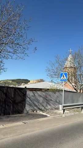 The video shows a drive-by view of a church and a fortress on a sunny day. Initially, the camera focuses on the church, showcasing its unique architecture, including a tall spire topped with a cross. As the vehicle moves, the view shifts to reveal an old stone fortress situated atop a hill in the distance. The fortress is partially obscured by trees and other buildings, suggesting a historical settlement in the area. The landscape is characterized by rolling hills and a clear blue sky, providing a serene backdrop to the historical structures. A road with a silver guardrail is visible in the foreground, indicating the viewer is traveling by vehicle.