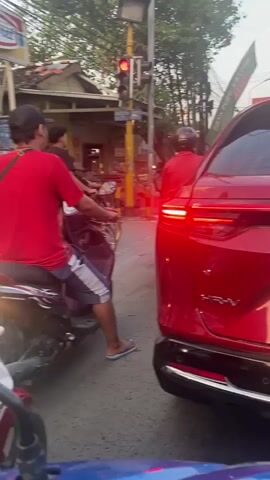 The video shows a street scene with traffic. A person wearing a red shirt and shorts is sitting on a motorcycle at a red traffic light. Another person in a red shirt is standing next to a red car. There are other motorcycles and people in the background. A convenience store sign is visible on the left side of the image. The traffic light is red.