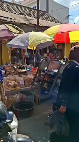 The video captures a bustling outdoor market scene. In the foreground, a person wearing a black headscarf and jacket walks away from the camera, while another person wearing a black helmet and jacket is partially visible, facing the first person. To the left, market stalls are set up with various goods displayed under colorful umbrellas. Eggs and packaged snacks are visible on a table. A trolley loaded with items is positioned in the center of the frame. Further into the market, a woman in a headscarf and patterned clothing sits by a stall selling produce, including what appear to be cucumbers. In the background, other market stalls line the street, some displaying colorful merchandise. An ATM sign is visible hanging above one of the stalls. The environment appears to be a sunny day with a partly cloudy sky.