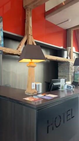 The video begins with a close-up of a hotel reception desk. The desk is dark wood with the word "HOTEL" carved into the front. There are two lamps on the desk, one on each side, with brown lampshades and decorative bases that resemble lighthouse towers. A white speaker is on the left side of the desk, and to its right is a computer monitor, a card reader, and some brochures. Behind the desk, a stone wall is visible, along with wooden beams and an orange wall.

The camera then pans to the right, revealing a large arched window with a view of a golf course. Two brown leather armchairs are positioned in front of the window, with a small black coffee table between them. The coffee table has magazines on it. Outside, a row of golf carts is parked, and a golf course with trees and rolling hills can be seen in the distance. The sky is cloudy, with the sun setting. A large green potted plant is to the right of the armchairs.

The camera continues to pan, showing more of the golf course and the sky. Finally, the video shows a glimpse of a pathway leading outside, paved with cobblestones, and bordered by stone walls and manicured bushes. The setting appears to be a golf resort or a similar leisure facility. There are no people clearly visible in the video.