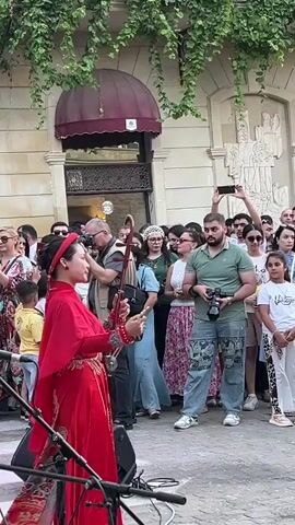 A woman dressed in a bright red traditional Vietnamese ao dai is playing a two-stringed bowed musical instrument, possibly a dan nguyet or dan tranh, on a street. She is standing in front of a crowd of people who are watching and taking pictures and videos with their phones. The woman is wearing a red headscarf and has her hair pulled back. She is holding a bow in her right hand and her left hand is on the strings of the instrument. She is performing outdoors in front of a building with an awning and ivy growing on it. The crowd is diverse in age and includes men, women, and children. Some people in the crowd are holding cameras and cell phones to record the performance.