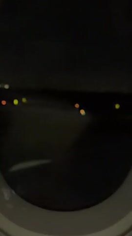 This video shows a view from an airplane window at night. The camera is focused on the runway lights, which are visible as small, colored dots in the darkness. The lights appear to be either yellow, orange, or blue. In the foreground, the edge of the airplane window is visible. The video is shaky, suggesting that the airplane is in motion. The image is also blurry, which may be due to the low light conditions or the movement of the camera.