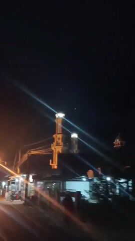 The video shows a nighttime street scene with a mosque minaret prominently lit. There are streetlights casting long streaks of light across the frame, and some vehicles are visible on the road, including a motorcycle, a truck, and a car. The scene appears to be in a populated area with buildings and some stalls lining the street. The camera is moving, giving a sense of passing by the scene.