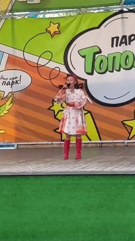 A young girl in a traditional Russian dress and red boots stands on a stage and sings into a microphone. The dress is white with red and gold embroidery. The stage is wooden and set up outdoors, with a green carpet in front of it. Behind the girl is a large banner with colorful graphics, including a comic book-style speech bubble that reads "Парк Тополя" (Park Topolya) in large green letters. Another speech bubble on the left says "Это наш парк!" (This is our park!). The banner has a bright orange and yellow striped background with green and yellow stars. The girl appears to be performing at a daytime event, possibly a festival or concert in a park. The overall atmosphere seems cheerful and festive.