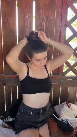 A young woman with brown hair pulled into a messy bun is in the process of styling her hair. She is wearing a black spaghetti-strap crop top and dark denim shorts. She is outdoors, sitting on a wooden bench in what appears to be a gazebo or outdoor covered seating area, with a rustic wooden wall behind her. Sunlight streams through the gaps in the wooden planks, indicating it's daytime. To her left, a black purse is visible, and to her right, a black hairbrush rests on a wooden surface, possibly a table. The woman's expression is focused on her task. The overall scene suggests a casual, warm-weather setting, likely in Drohovyzh, Ukraine. There is no visible text in the video.