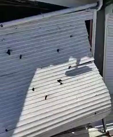 The video shows the damaged metal shutter of a store. The shutter has numerous puncture marks and is partially collapsed, revealing the interior of the store, which appears to have shelves stocked with various items. The lighting suggests it is daytime. No people are visible.