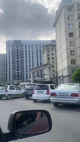 The video is a first-person perspective shot from inside a car. The view is primarily out the side window and rearview mirror. The car is in traffic, stopped on a paved road. In front of the car are several other vehicles, including a white compact car, a dark colored SUV, and a white hatchback with a roof rack. Further ahead, there are two large, modern apartment buildings. The building on the right is a light beige color with many windows and a decorative top. The building on the left is taller and more grey in color with a grid-like pattern of windows. Between the buildings is an ornate entrance archway. The sky is cloudy and overcast. The overall impression is of an urban street scene with moderate traffic in front of upscale residential buildings.