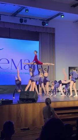 A group of young dancers performs on a stage for an audience. The dancers wear dark blue dresses with light blue accents. One dancer in the center wears a red leotard. The dancers on stage stand in two rows. In the background, a large projector screen is illuminated with blue light and text. The stage is dark, with spotlights highlighting the performers. A plume of white smoke rises from the left side of the stage. Members of the audience are visible in the foreground. A speaker stands at the front left of the stage.