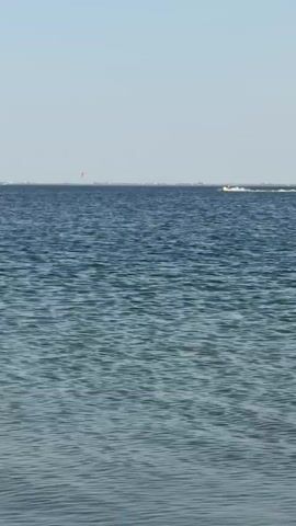 This media file captures a serene scene on the Black Sea near Ukrainka, Ukraine. The dominant subject is the vast, calm expanse of the dark blue sea, reflecting the clear, pale blue sky. Gentle ripples create a textured surface on the water.

In the distance, two distinct activities are occurring. On the right side of the frame, a white personal watercraft, likely a jet ski, is moving at speed, leaving a visible white wake. A person in yellow clothing is riding it, suggesting a recreational activity. To the far left, a small red kite is visible against the horizon, hinting at kiteboarding or a similar wind-powered sport. The distant shoreline is lined with what appear to be buildings and trees, providing a subtle indication of land.

The time of day appears to be late morning or early afternoon, given the bright, even lighting and the clear sky, which suggests fair weather. There are no visible signs of clouds. The overall mood conveyed is one of peacefulness and outdoor recreation, with the two distant activities adding a touch of dynamic energy to the tranquil setting. No text is visible in the media file.