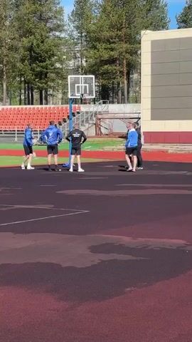 Several people are gathered near a basketball hoop on an outdoor court. The court appears to be a dark maroon or brown color, and there are some dark, wet patches on the surface. An athletic field with red seating is visible in the background. The people are a mix of genders and are wearing casual athletic clothing; some are in blue jackets. Some individuals are standing with their hands on their hips, observing, while others seem to be shooting a basketball. The environment includes trees, a building with gray and red walls, and a clear sky.