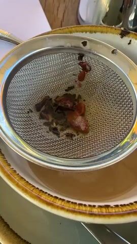 A close-up video shows a sieve with tea leaves being held over a cup of tea. The tea leaves are dark brown and appear to be in a wet clump. The tea in the cup is a light brown color with some foam on top. The cup is white with a brown rim and is sitting on a saucer. A spoon is visible in the saucer. The background appears to be a wooden table. The video is shot from above.