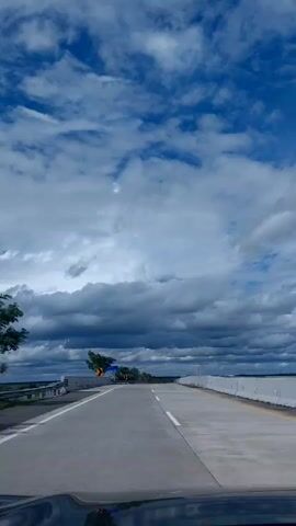The view from the windshield of a car driving on a highway on a partly cloudy day shows a clear road ahead with concrete barriers on both sides. The sky is filled with a mix of blue sky and white and gray clouds. Road signs with arrows point to the side of the road. There are a few trees and green areas visible in the distance. The car is moving forward at a steady pace.