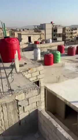 The video shows a rooftop in what appears to be a Middle Eastern city. Several red, white, and green water tanks are scattered across the flat surface, along with a satellite dish. A man dressed in white can be seen in the background talking on a mobile phone. The rooftop is made of concrete blocks, and there are other buildings visible in the distance. The sky is clear and the lighting indicates it is daytime.