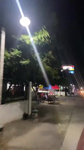 The video is shot at night in an outdoor environment, likely a street in a commercial area. The camera is moving, suggesting it is being recorded from a vehicle or while walking.

In the foreground, a tree with green leaves partially obstructs the view. Beneath the tree, there is a street with a paved surface. A small food cart or stall is visible, covered by colorful umbrellas (red, yellow, and blue). A motorcycle is parked next to the stall. There are some plastic chairs on the ground.

Further down the street, a brightly lit sign is visible, appearing to be for a convenience store, possibly "Indomaret" based on the colors and logo. There are streetlights illuminating the area. The sign has multiple panels, including one with text that might indicate services like restrooms.

The background shows buildings and structures that appear to be shops or houses. Some lights are on in these buildings. A few more motorcycles and possibly a car are parked along the street. The overall atmosphere is that of a typical street scene at night in a populated area, with commercial activity present. The lighting is a mix of streetlights and illuminated signs, creating a nocturnal ambiance.