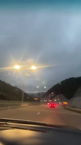 The video is a first-person perspective from inside a car traveling on a highway at dusk or early evening. Streetlights illuminate the road, and the sky is overcast. Several cars are visible on the highway, with their headlights and taillights on. The car the camera is in is moving forward, and the dashboard is partially visible in the foreground. The road is flanked by hills or mountains on either side. The overall atmosphere is somewhat moody due to the lighting conditions and the overcast sky.