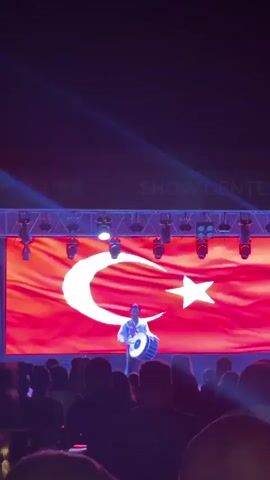 A man stands on a stage in front of a large projection of the Turkish flag. He is wearing a white shirt and a blue hat. He is playing a large drum with two drumsticks. There is a crowd of people in the foreground, silhouetted against the stage lights. The stage is equipped with a lighting rig. Text that appears to read "SHOW CENTER" is visible above the stage. The overall lighting is dark with bright spotlights.