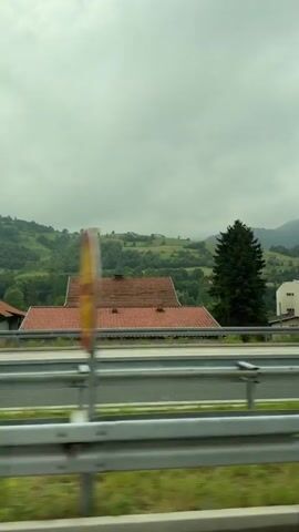 A side view from a vehicle shows a landscape of rolling green hills under a cloudy sky. In the foreground, a highway guardrail is visible. Beyond the guardrail, there are several houses with red-tiled roofs and some larger buildings, including a white multi-story structure. Evergreen trees are scattered throughout the scene, with a dense cluster of them on the right side. The hills in the background are covered in lush green vegetation and appear to be forested in places. The overall impression is of a rural or semi-rural setting.