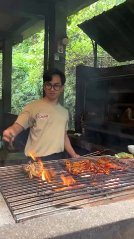A young man is grilling food. He is wearing glasses and a tan T-shirt with a logo on the upper-left side. The shirt has the word “Cupit” and the words “BBQ Grill” written under it in small font. He is using tongs to turn chicken that is cooking on a grill with a blazing fire underneath it. To the right of the chicken is an assortment of other food being grilled. Behind the man is a large black grill that looks like a smoker. The man is in an open outdoor area that is covered by a green-and-brown roof. Green trees and bushes are visible in the background.
