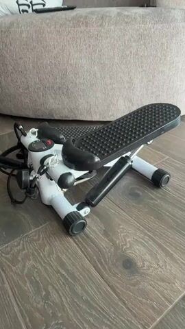 The video shows a white and black mini stepper machine resting on a wooden floor. The machine is in the foreground, with a large, neutral-colored couch or ottoman in the background. The stepper has two pedals, which appear to be made of black textured material. There is a digital display on the front of the machine, showing some readings. Wires and a handle are also attached to the machine. The lighting is natural, and the scene appears to be indoors, possibly in a living room. The camera is positioned at a low angle, looking down at the stepper. The video is brief, showing only slight movement or angles of the stepper.