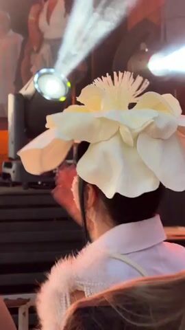 The video shows a close-up of a person from behind, wearing a large, white, flower-shaped hat and a white jacket with white fur trim and bejeweled embellishments on the shoulder. The person's dark hair is visible beneath the hat. Another person with reddish-brown hair is visible in the foreground to the right. In the background, stage lights are illuminating a dark area, suggesting a performance or event setting. The lighting is dramatic, with bright beams of light cutting through the darkness. The video appears to be shot from a handheld perspective, with some movement and blur. The overall impression is of a festive or celebratory atmosphere.