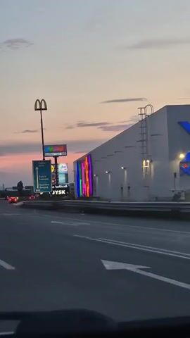 The video is a first-person perspective, likely from inside a vehicle, looking towards a roadside scene at dusk. The sky displays soft pastel colors of pink, orange, and purple from the setting sun, with scattered clouds. To the left, a tall pole supports a McDonald's golden arches sign and other illuminated advertisements for businesses such as Jumbo, Lidl, dm, Paolo Botticelli, and JYSK. A directional sign points towards Sofia, Pernik, and Belgrade. A large, white building with colorful neon lettering spelling out "JUMBO" is prominently displayed on the right. The road in the foreground shows lane markings and a distant traffic light. Cars are visible moving in the distance. The overall impression is of a commercial area on the outskirts of a city as evening approaches.