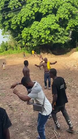 The video captures an outdoor scene in Unwana, Nigeria, depicting several men engaged in what appears to be a traditional dance or performance on a dusty, earthy ground. In the foreground, a man wearing a white polo shirt, blue jeans, and carrying a grey crossbody bag observes the activity. Behind him, another man wears a black t-shirt with "UJU MEMORIAL UNIVERSITY" visible text.

Further back, several shirtless men are actively participating. They move rhythmically, some adopting stances reminiscent of sparring or a ceremonial fight, extending their arms and bending their bodies. One man wears a bright yellow cloth draped over his shoulders. Children are also scattered in the background, some observing, one running.

The setting is characterized by a large, lush green tree dominating the background, suggesting a rural or village environment. The ground is uneven and appears to be bare earth. The activity takes place during daytime, under bright, natural light. The overall mood appears to be one of cultural celebration or ritual.