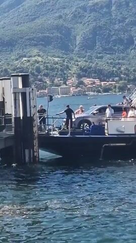 A car is being loaded onto a ferry. People are standing on the ferry and on the dock. The ferry is on a body of water, with mountains and buildings in the background.