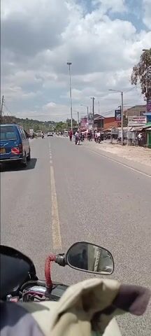 The video depicts a view from a moving motorcycle traveling down a relatively busy street in an urban or semi-urban area. There are various cars, motorcycles, and pedestrians visible along the road and sidewalks. The road is paved with a solid yellow line dividing traffic.  Buildings line the sides of the street, with signs indicating businesses, including what appears to be a high school and a bank. A tall light pole stands prominently on one side of the road. The sky is partly cloudy, suggesting daytime. The camera is pointed forward from the driver's perspective, capturing the road ahead and reflections in the motorcycle's side mirror. The overall impression is of a normal day in a moderately active commercial area.