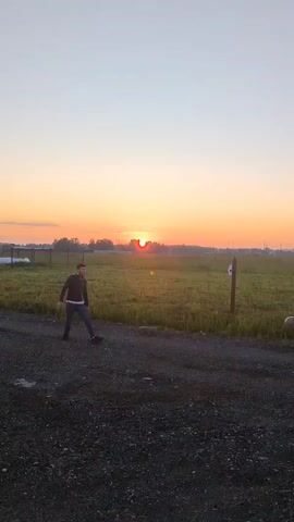 Two young men are playing catch with a soccer ball in an open field at sunset. The sun is low on the horizon, casting a warm glow over the scene. The man on the left is wearing a dark jacket and jeans, while the man on the right is wearing a black shirt and pants. The ball is in mid-air, having been thrown by the man on the left. The field is covered in tall grass, and there are some trees and buildings in the distance. The ground in the foreground is covered with gravel.