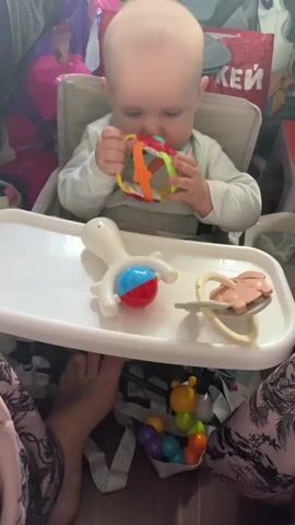 A baby is sitting in a high chair and playing with a rattle. The baby is holding the rattle in both hands and shaking it. There are other toys on the tray of the high chair, including a white toy with a red and blue ball, a pacifier, and a caterpillar-shaped toy with colorful balls. The background is cluttered with bags and other items. The lighting is dim.