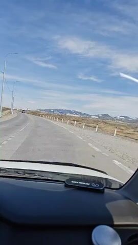 A first-person perspective video captures a car driving on a paved road under a bright blue sky with scattered white clouds. The road is mostly clear, with a few white posts lining the right side. In the distance, there are buildings and hills, some of which appear to be snow-capped. The dashboard of the car is visible in the foreground, with a small black device with a logo on it. The overall impression is of a daytime journey through a somewhat barren landscape.
