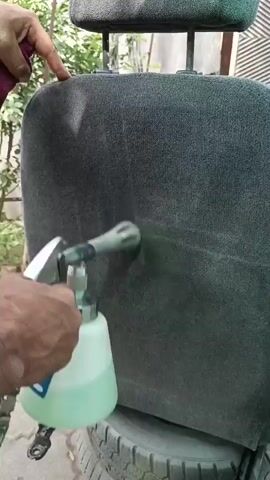 A person's hands are holding a cleaning device that is spraying a green liquid onto a car seat. The seat is dark gray and appears to be fabric. The cleaning device has a silver nozzle and a clear plastic bottle containing the green liquid. The person's left hand is holding the top of the seat, while their right hand is operating the cleaning device. The background is out of focus but appears to be outdoors, with some greenery and a wall visible. The seat is resting on what appears to be a tire.