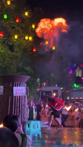 At night, a crowd gathers around a large, ornate fire pit constructed from bamboo. The area is illuminated by numerous colorful lanterns strung from trees overhead and by handheld torches carried by some audience members. In the center of the scene, a man dressed in traditional clothing, including a red vest, is performing a fire-eating or fire-dancing ritual. He is using torches to interact with the fire and the large pit. Some members of the audience are recording the performance with their phones. A sign on the bamboo structure, written in Chinese characters, appears to be a warning or instruction related to the fire. The overall atmosphere suggests a cultural festival or event.