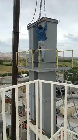 A construction worker in a blue denim jacket and jeans is working on a tall industrial structure. He is on a small platform, reaching up to attach chains to a large, gray, rectangular metal component that is suspended from above by a crane. The worker is positioned with his back to the camera, arms raised. The industrial structure appears to be a tall, multi-sectioned tower made of gray metal. In the background, there is a landscape with rolling hills and some vegetation. The sky is overcast.