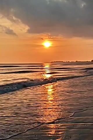 The video shows a beach scene at sunset. The sun is setting over the ocean, creating a golden glow on the water and sky. Waves are gently lapping onto the shore. There is a distant landmass visible on the horizon. The sky is partly cloudy, with dark clouds overhead.