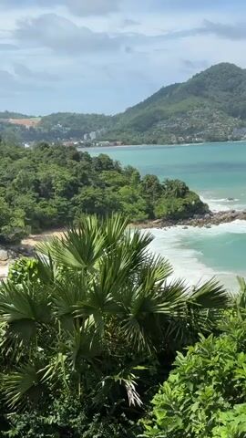 The video shows a scenic view of a tropical coastline. In the foreground, lush green foliage, including palm fronds, obscures the bottom portion of the frame. Beyond the foliage, a turquoise ocean with white-capped waves crashes against rocky outcrops and a sandy beach. In the distance, a verdant, tree-covered mountain rises, dotted with buildings along its slopes. The sky is partly cloudy, with hints of blue visible. The overall impression is of a tranquil, natural landscape. There are no people or specific actions depicted.
